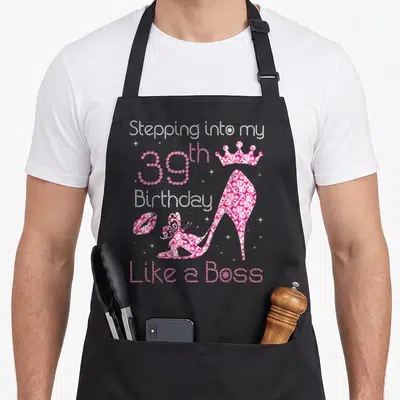 39th bbq boss apron - Pink high heel shoe with crown and lips surrounded by Stepping into my 39th Birthday Like a Boss — shown on a black cotton canvas apron against a white background