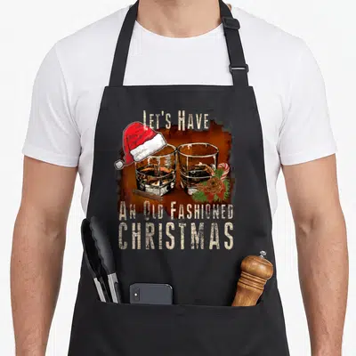 Drinker old fashioned aprons - Two whiskey glasses with Santa hat and cinnamon stick, distressed text — shown on a black cotton canvas apron against a white background