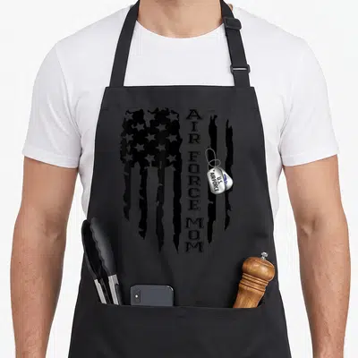 Flag call mom apron - Distressed black flag with vertical AIR FORCE MOM text and silver dog tags — shown on a black cotton canvas apron against a white background