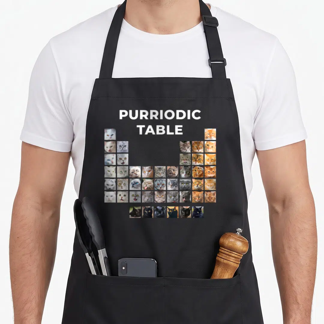 Cat chemistry apron - Purriodic Table grid of cat faces in white, grey, orange, and black photos — shown on a black cotton canvas apron against a white background