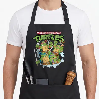 Break teenage mutant ninja apron - Four turtles with colored masks emerging from a white jagged tear below stacked — shown on a black cotton canvas apron against a white background