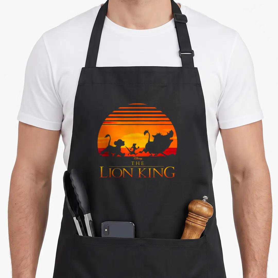 Sunset lion apron - Retro Lion King silhouettes running against orange striped sunset with stacked title text — shown on a black cotton canvas apron against a white background