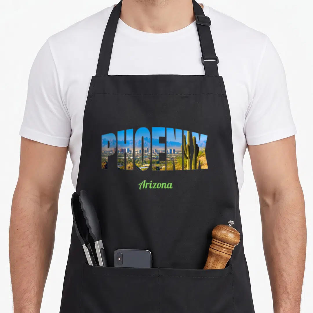 Phoenix sightseeing apron - PHOENIX text filled with city and mountain landscape photo with Arizona script below — shown on a black cotton canvas apron against a white background