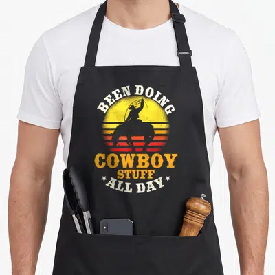 Doing farm apron - Retro cowboy silhouette on horseback with lasso against striped sun and text BEEN DOING COWBOY STUFF — shown on a black cotton canvas apron against a white background