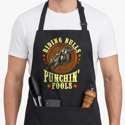Bull riding quotes apron - Riding Bulls Punchin Fools text with realistic bull rider in orange circle — shown on a black cotton canvas apron against a white background