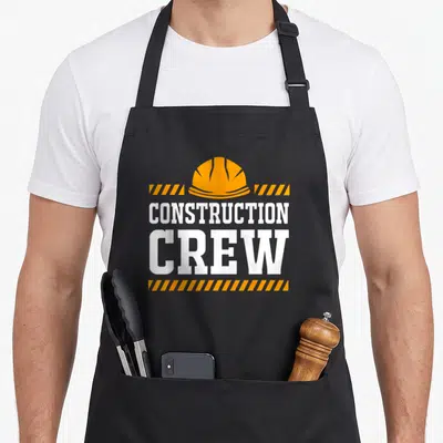 Family construction apron - Orange hard hat with white CONSTRUCTION CREW text and orange hazard stripes — shown on a black cotton canvas apron against a white background