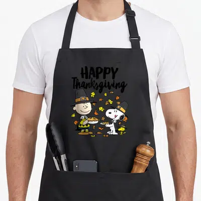 Charlie thanksgiving apron - Charlie Brown and Snoopy in pilgrim hats holding pies with scattered yellow and orange — shown on a black cotton canvas apron against a white background