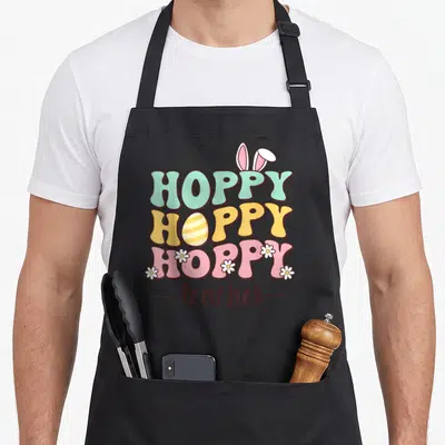 Teacher egg easter bunny aprons - Stacked hoppy text with bunny ears, striped egg, and daisies above teacher script — shown on a black cotton canvas apron against a white background