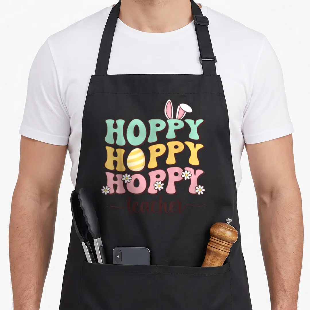 Teacher egg easter bunny aprons - Stacked hoppy text with bunny ears, striped egg, and daisies above teacher script — shown on a black cotton canvas apron against a white background