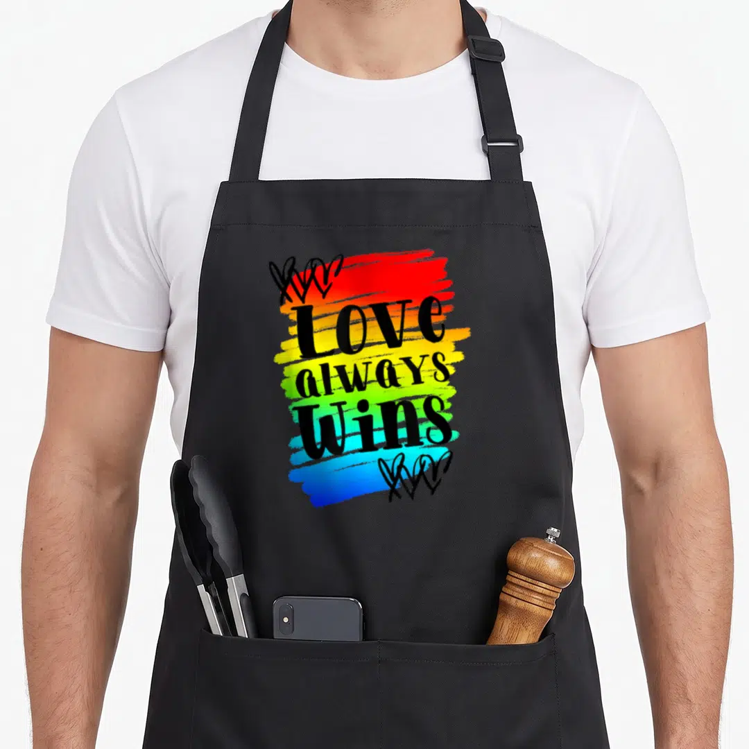 Alway gay pride apron - Love Always Wins text over rainbow brushstroke with black heart clusters — shown on a black cotton canvas apron against a white background