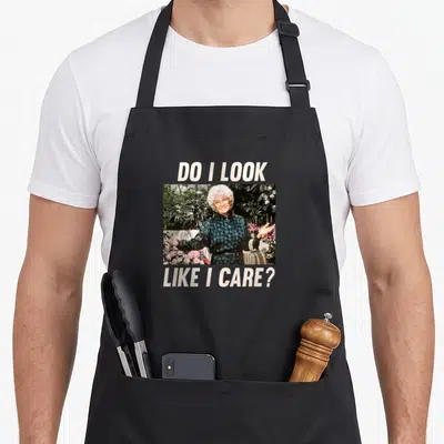 Sophia golden apron - Photo of woman shrugging with text DO I LOOK LIKE I CARE above and below — shown on a black cotton canvas apron against a white background