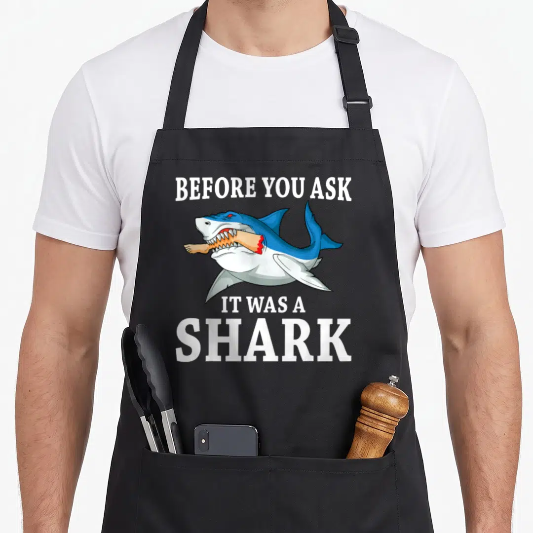 Before leg apron - Cartoon blue shark biting a peach foot with white text BEFORE YOU ASK IT WAS A SHARK — shown on a black cotton canvas apron against a white background