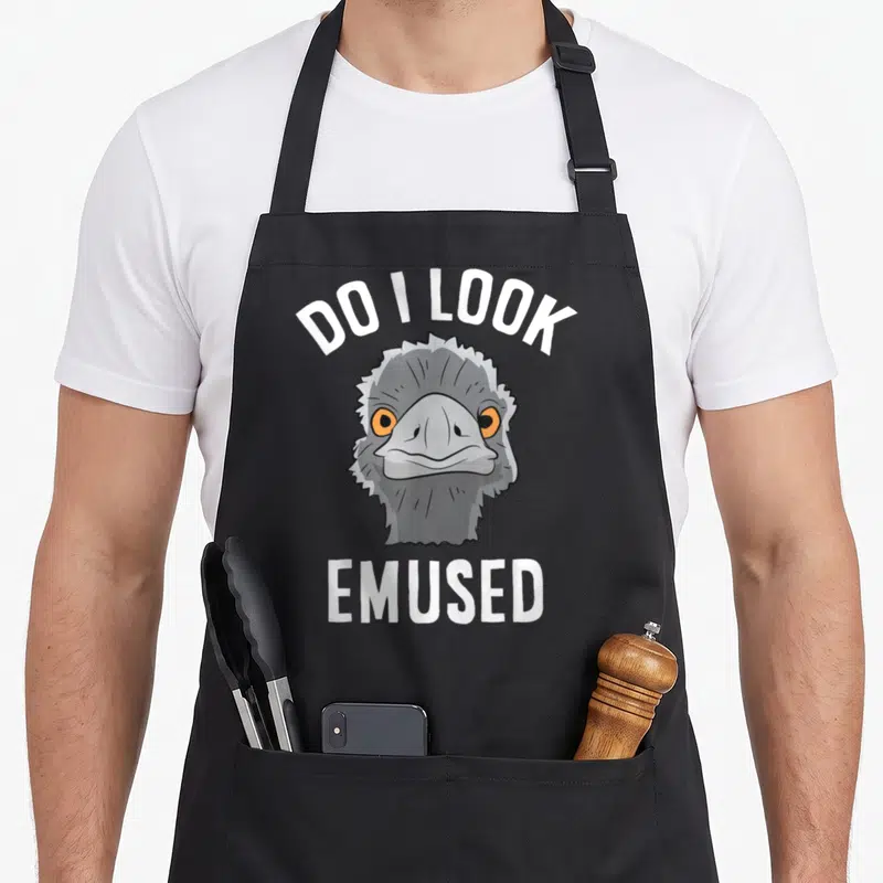 Amused animal apron - Cartoon emu head with orange eyes and white text DO I LOOK EMUSED arched above and straight below — shown on a black cotton canvas apron against a white background