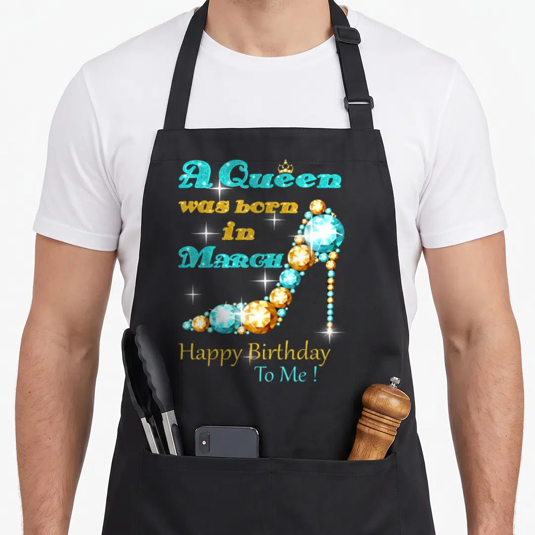 March baking queen apron - A Queen born in March text with gem high heel shoe in blue and gold — shown on a black cotton canvas apron against a white background