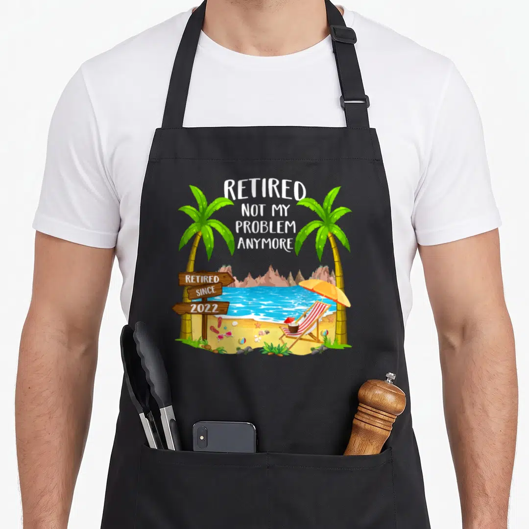 Beach retirement apron - Cartoon beach scene with palm trees, lounge chair, and text retired since 2022 on wooden sign — shown on a black cotton canvas apron against a white background