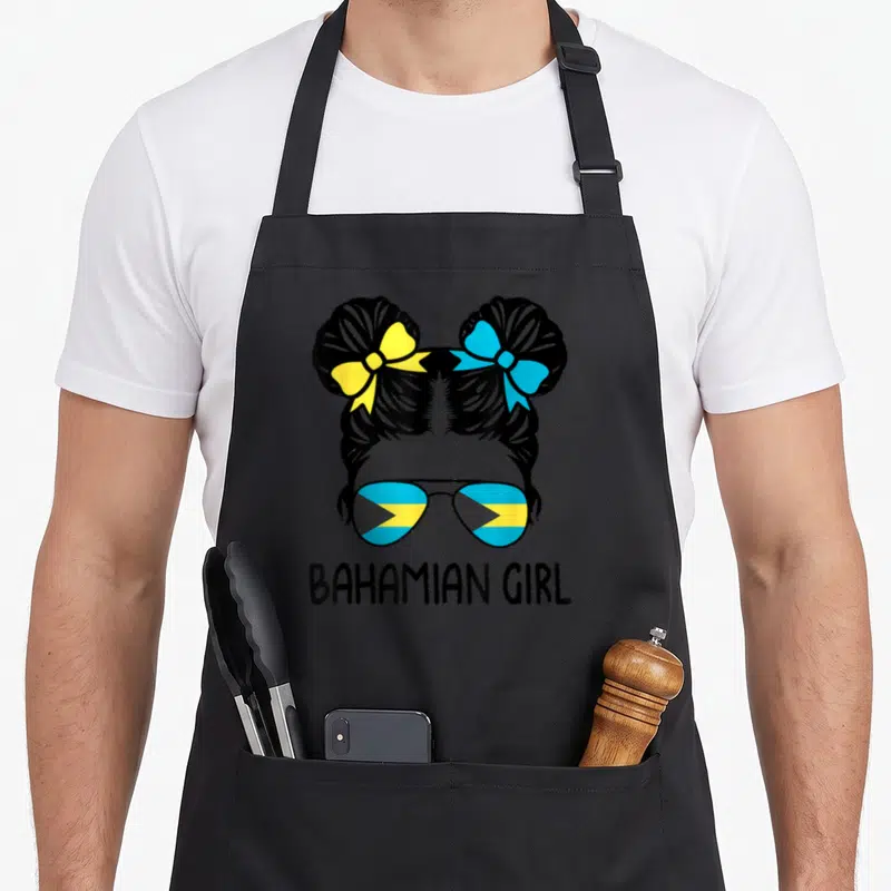 Bahamian messy aprons - Girl silhouette with two buns, yellow and blue bows, flag sunglasses, BAHAMIAN GIRL text — shown on a black cotton canvas apron against a white background