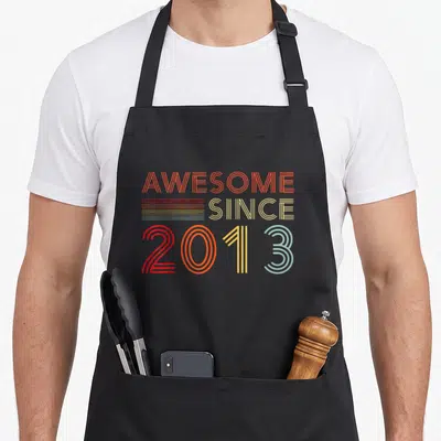 13yr birthday apron - Stacked AWESOME SINCE 2013 text in orange yellow red and light blue retro lines — shown on a black cotton canvas apron against a white background