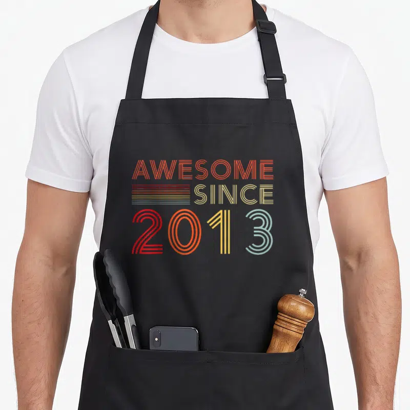 13yr birthday apron - Stacked AWESOME SINCE 2013 text in orange yellow red and light blue retro lines — shown on a black cotton canvas apron against a white background