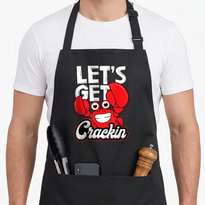 Crackin crab apron - Red cartoon crab with white text LET'S GET Crackin and black outline — shown on a black cotton canvas apron against a white background