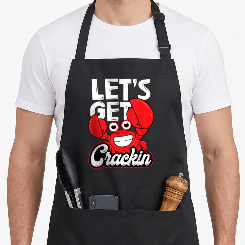 Crackin crab apron - Red cartoon crab with white text LET'S GET Crackin and black outline — shown on a black cotton canvas apron against a white background