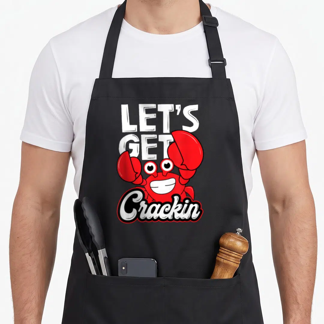 Crackin crab apron - Red cartoon crab with white text LET'S GET Crackin and black outline — shown on a black cotton canvas apron against a white background