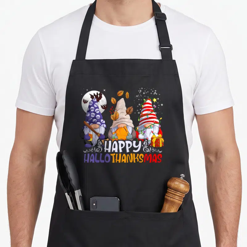 Hallothanksmas christmas gnome apron - Three gnomes with skull hat, pumpkin, and lights under Happy Hallothanksmas text — shown on a black cotton canvas apron against a white background