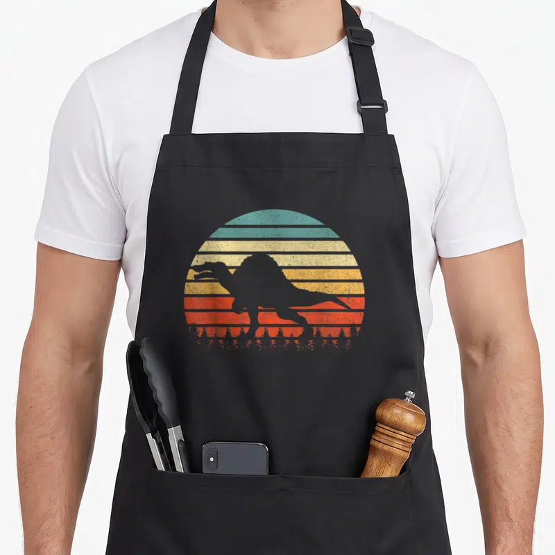 Dinosaurus spinosaurus apron - Grey Godzilla silhouette over retro striped sun in teal yellow orange and red — shown on a black cotton canvas apron against a white background