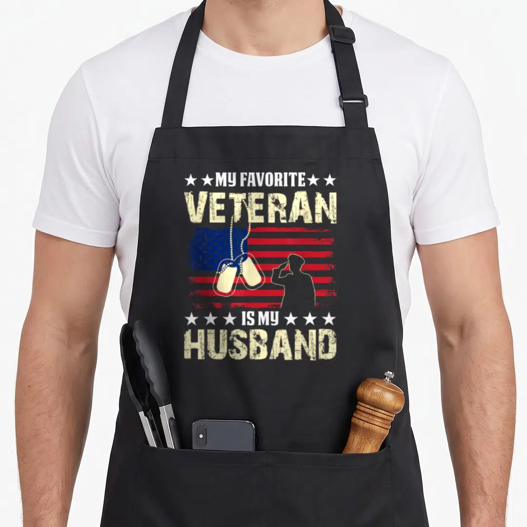 Patriot best husband apron - My Favorite Veteran Is My Husband text with saluting soldier, dog tags, and distressed flag — shown on a black cotton canvas apron against a white background
