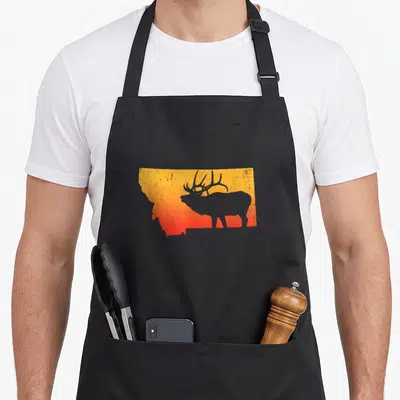 Elk outdoor apron - Minimalist elk silhouette inside Montana state map shape with orange and red gradient background — shown on a black cotton canvas apron against a white background