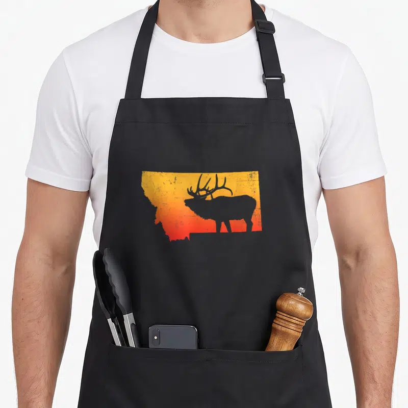 Elk outdoor apron - Minimalist elk silhouette inside Montana state map shape with orange and red gradient background — shown on a black cotton canvas apron against a white background
