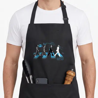 Street road apron - Four men walking on blue crosswalk with The Beatles Abbey Road text — shown on a black cotton canvas apron against a white background