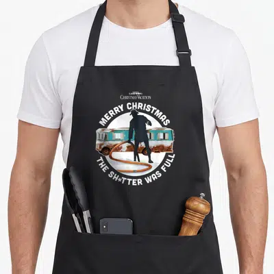 Shitter funny animals vacation and holidays apron - Man with hose in front of blue RV with Merry Christmas text — shown on a black cotton canvas apron against a white background