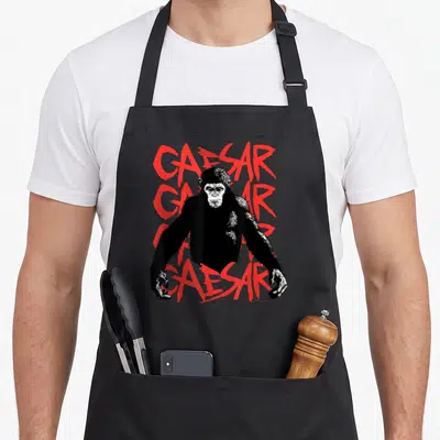 Planet ape apron - Black and white chimpanzee standing in front of four stacked red CAESAR words in distressed font — shown on a black cotton canvas apron against a white background