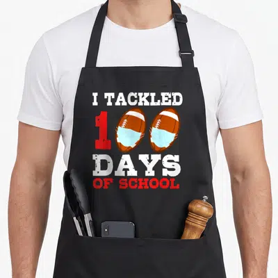 Football teacher apron - I TACKLED 100 DAYS OF SCHOOL text with masked footballs forming zeros — shown on a black cotton canvas apron against a white background
