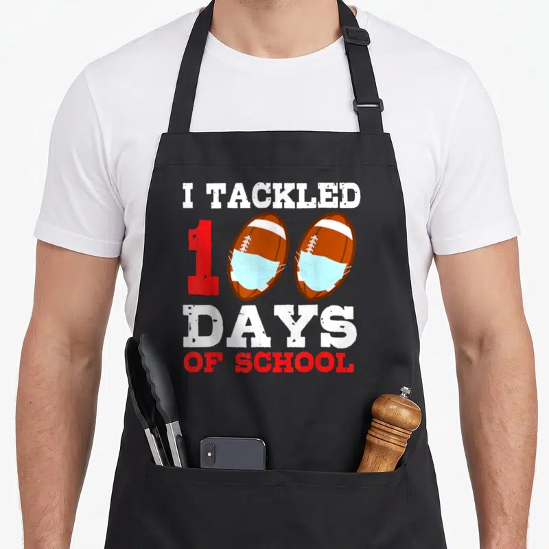 Football teacher apron - I TACKLED 100 DAYS OF SCHOOL text with masked footballs forming zeros — shown on a black cotton canvas apron against a white background