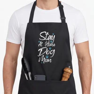 Stay dog at home aprons - Stacked white script text reading Stay At Home Dog Mom with scattered light blue paw prints — shown on a black cotton canvas apron against a white background