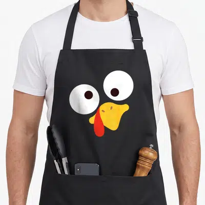 Pilgrim turkey apron - Cartoon turkey face with big white eyes, yellow beak, and red wattle — shown on a black cotton canvas apron against a white background