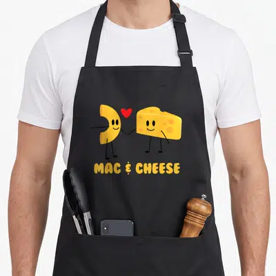 Mac veggie apron - Cartoon macaroni and cheese wedge holding hands with red heart and yellow text — shown on a black cotton canvas apron against a white background