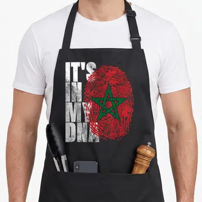Dna african apron - IT'S IN MY DNA text over red fingerprint with green star — shown on a black cotton canvas apron against a white background