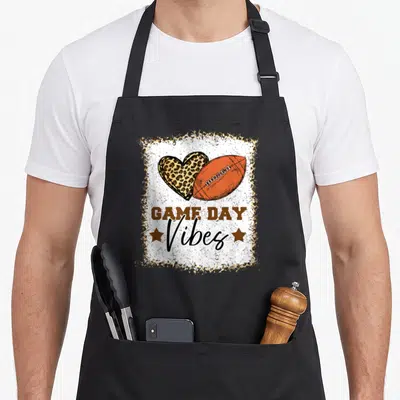 Game holiday football apron - Distressed GAME DAY Vibes text below leopard heart and orange football in brown frame — shown on a black cotton canvas apron against a white background
