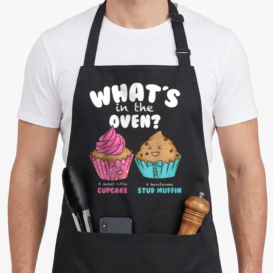 Reveal muffin man apron - Cartoon cupcake and muffin with What's in the Oven pun text — shown on a black cotton canvas apron against a white background