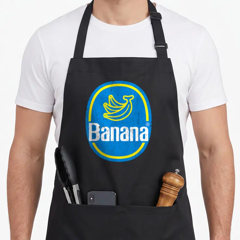 Banana funny halloween aprons - Blue oval with yellow border, yellow banana bunch, white 'Banana' text, retro style — shown on a black cotton canvas apron against a white background