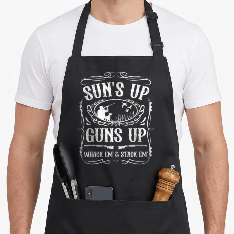Sun grill hunter apron - White distressed text Suns Up Guns Up with hunter silhouette, dog, flying ducks, and two — shown on a black cotton canvas apron against a white background