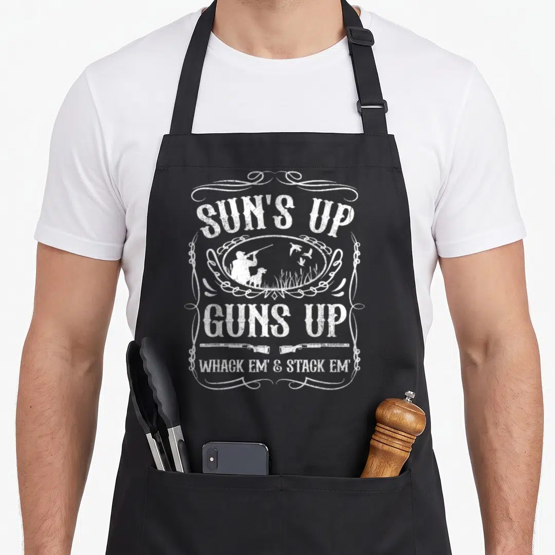 Sun grill hunter apron - White distressed text Suns Up Guns Up with hunter silhouette, dog, flying ducks, and two — shown on a black cotton canvas apron against a white background