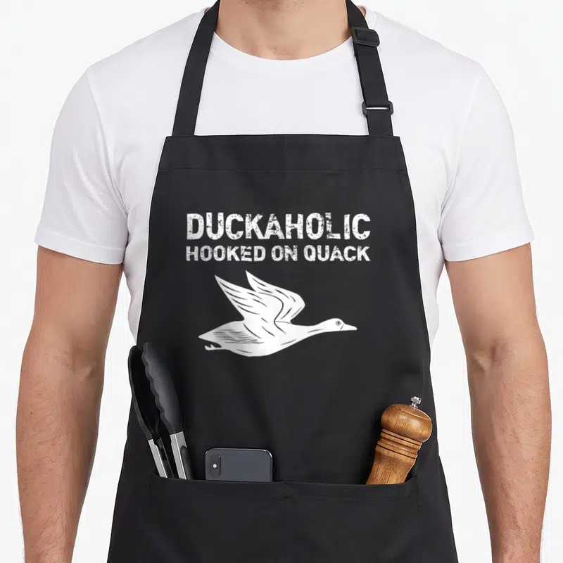 Duckaholic hunter apron - White distressed text reading Duckaholic Hooked On Quack above a flying duck illustration — shown on a black cotton canvas apron against a white background