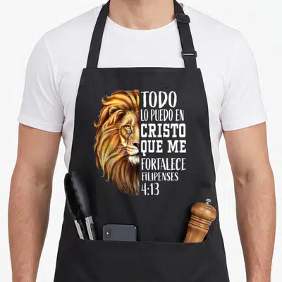 Spanish lions apron - Realistic lion head next to stacked Spanish text TODO LO PUEDO EN CRISTO FILIPENSES 4:13 in white — shown on a black cotton canvas apron against a white background