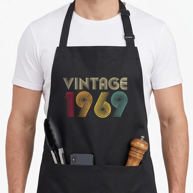 57th birthday apron - VINTAGE 1969 stacked text in parallel lines colored beige, red, orange, yellow, and teal — shown on a black cotton canvas apron against a white background