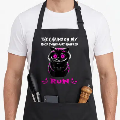 Chain cat apron - Black cartoon cat with pink eyes and text The Chains On My Mood Swing Just Snapped Run — shown on a black cotton canvas apron against a white background