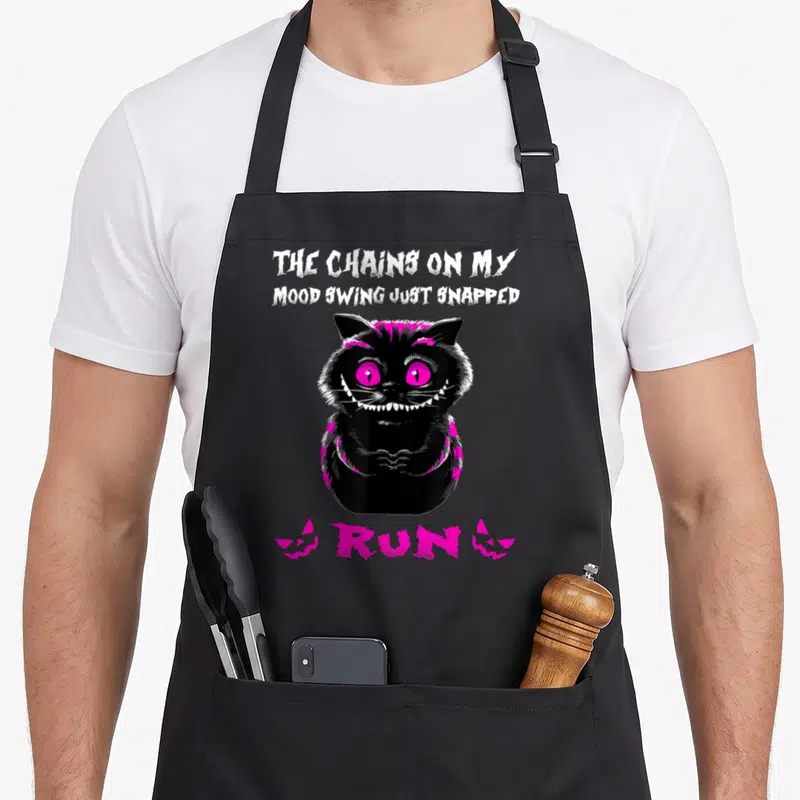 Chain cat apron - Black cartoon cat with pink eyes and text The Chains On My Mood Swing Just Snapped Run — shown on a black cotton canvas apron against a white background