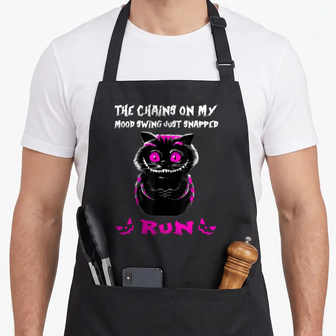 Chain cat apron - Black cartoon cat with pink eyes and text The Chains On My Mood Swing Just Snapped Run — shown on a black cotton canvas apron against a white background